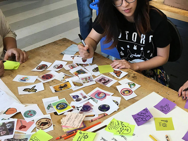 Woman pulls inspiration from a table full of printed graphic designs during a Design Bloc workshop on creating patches.
