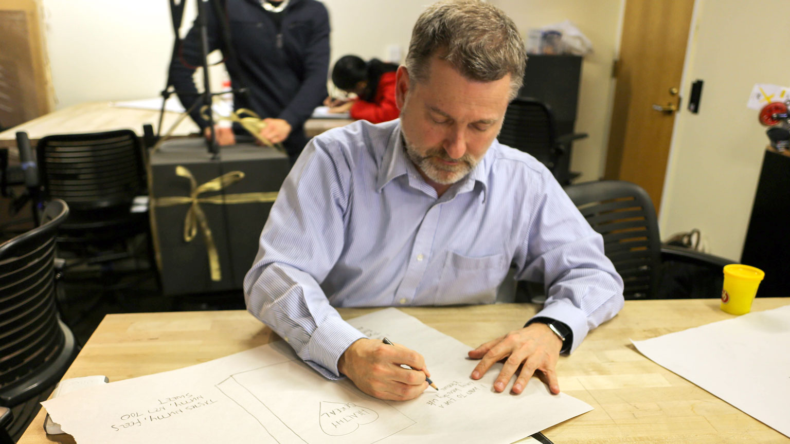 A man sketches in a large notebook while a cameraman films from behind, photo taken during a Design Bloc activity on narrative storytelling. 
